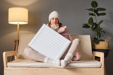 Seasonal temperature changes. Getting warm in winter. Calm woman wearing cap and coat sitting on couch and embracing heater battery trying to get warm.