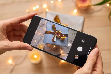 Hands holding phone and taking photo of Christmas gift boxes, lights on wooden background and candles stylish festive photography.