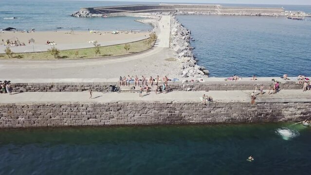 Aerial shot travelling along stone jetty with swimmers coming out of the water up stone steps in Mutriku, Spain.