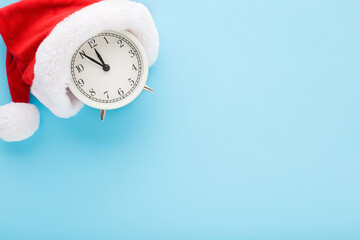 Red hat with white fur and alarm clock on blue table background. Pastel color. Closeup. Countdown...