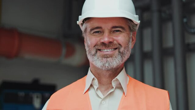 Close up portrait of aged construction manager dressed in orange vest and white hard hat posing at workplace. Smiling bearded male expressing positive emotions and looking straight at manufactory.