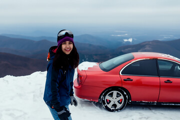 girl on the mountain with a car. a girl in winter clothes and mountain glasses stands on a snowy mountain near a red car