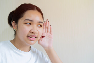 A young attractive Asian girl in a white t-shirt holding her palm near her ear and listening to gossip.