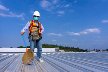 Worker wearing full safety body harness holding a broom in hand  working on roof top for cleaning metal roof sheet - obrazy, fototapety, plakaty