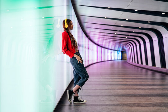 Thoughtful woman with headphones leaning on illuminated wall in underground tunnel