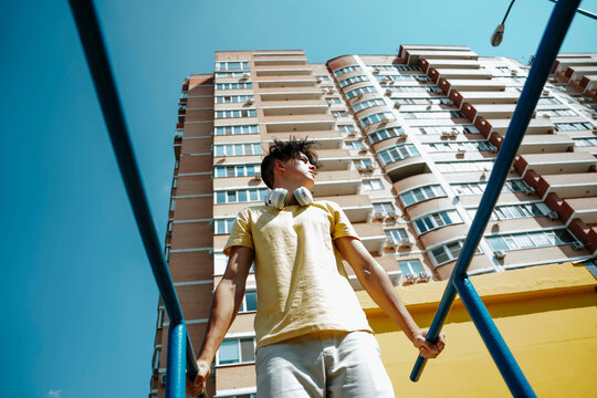 Young man with wireless headphones standing near bars in front of building