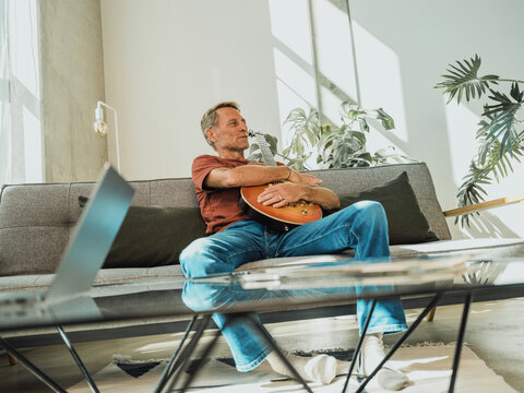 Man with guitar sitting on sofa at home - Powered by Adobe