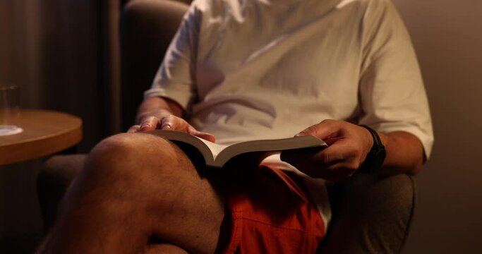 Relaxed man reads favourite book in armchair in hotel lounge in evening. Male tourist enjoys reading fascinating edition on holiday abroad slow motion