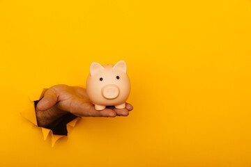 Hand with a piggy bank through a yellow paper hole with copy space