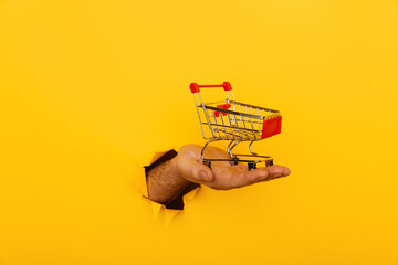 Male hand holds through a hole a mini grocery shopping trolley on a yellow paper background