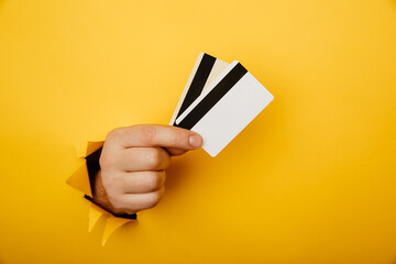 Male hand is holding credit bank cards in torn hole of yellow background. Online shopping, purchasing products concept.
