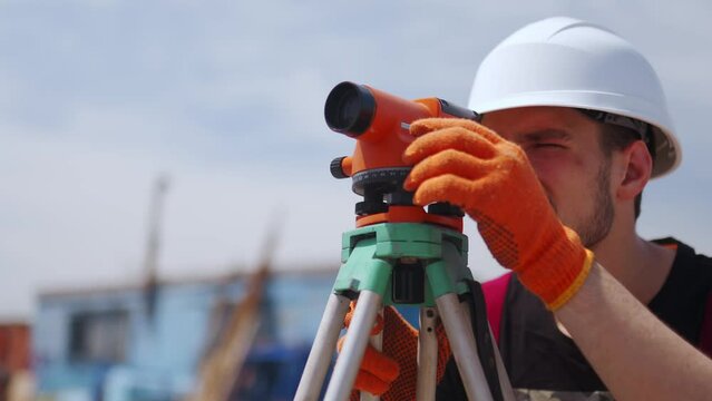 Construction worker uses optical level on construction site. Civil engineer with a theodolite doing land surveying work. Man wearing hard hat and work gloves measures angles for building plans.
