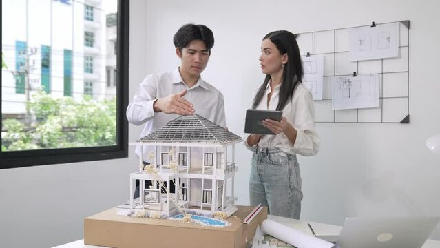 Beautiful young caucasian project manager brief asian engineer by using tablet while engineer measuring house model on meeting table with blueprint, architectural equipment scatter around. Immaculate