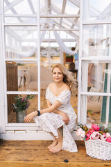 Vertical portrait of happy blonde young woman in beautiful dress sitting in doorstep of summer gazebo house on sunny day smiling looking at camera. Concept of summer vacation
