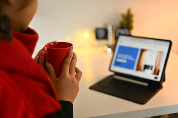 Young woman drinking hot chocolate and browsing internet on digital tablet at cozy living room