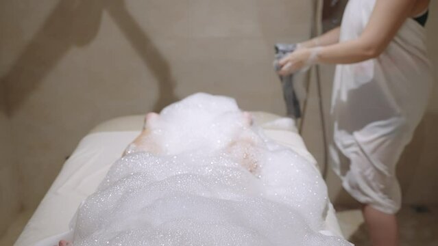 Spa worker washes woman under bubble soap foam with a stream of water in hammam shower. Female relax, gets body skin treatment, cleansing, wash. Anti aging and anti cellulite therapy in sauna.