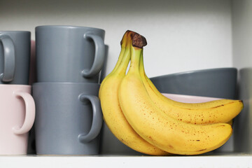 Bananas in kitchen cabinet. Coffee mugs. Tea cups. Milk soup bowls. Yellow fruits in kitchen. Open door kitchen cabinet with hidden bananas inside. Ripe bananas with small brown stains. - obrazy, fototapety, plakaty