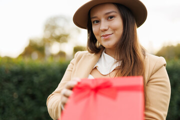 Portrait of charming lady in beige classic coat holding present box in autumn park at sunset.