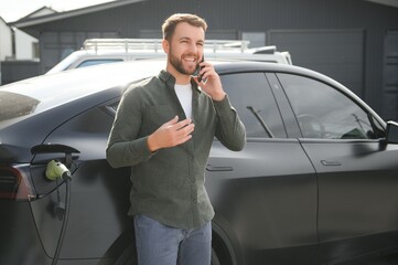 Happy man using smart phone and charging car at vehicle charging station