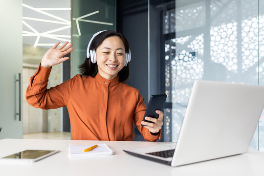 Young woman inside office at workplace listening to relaxing soothing music, Asian businesswoman in headphones using app on phone, sitting with laptop at table, wearing headphones.