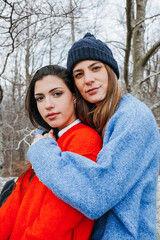 Young beautiful lesbian couple in bright knitted sweaters embracing in the forest, looking at camera.