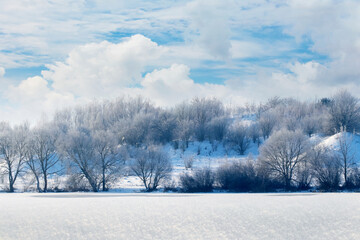 Winter landscape with trees on the banks of the river and a picturesque blue sky with white curly clouds in sunny weather