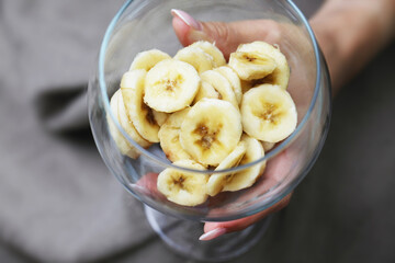 Top view banana salad. Wine glass in hand with fruit slices inside. Banana slices in glass. Woman hand holding fruit slices. Transparent bowl with banana pieces.