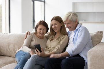 Cheerful elder couple of parents and adult child woman using mobile phone together, taking family selfie at home, sitting on sofa, posing, smiling, talking on video call on smartphone - obrazy, fototapety, plakaty