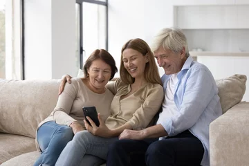Fotobehang Cheerful elder couple of parents and adult child woman using mobile phone together, taking family selfie at home, sitting on sofa, posing, smiling, talking on video call on smartphone © fizkes