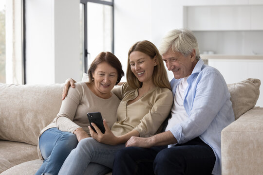 Cheerful elder couple of parents and adult child woman using mobile phone together, taking family selfie at home, sitting on sofa, posing, smiling, talking on video call on smartphone