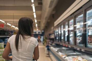 Young woman in the freezer section of the supermarket. Middle-aged girl with her back turned looking at the display cases of frozen products and pushing the cart. Batch cooking concept.