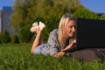 Portrait of a young smiling blonde woman lying on her stomach on the grass in the park with a laptop on a sunny day. Student communicates, studies with a laptop in a city park