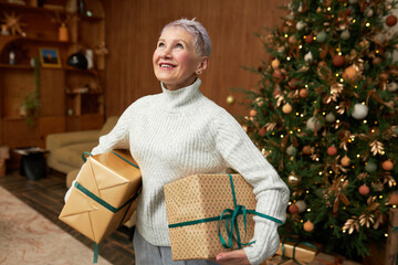 Happy overjoyed senior female in gray jumper holding big wrapped present boxes in both hands looking up thanking god for generosity and making her dreams come true, standing against christmas tree
