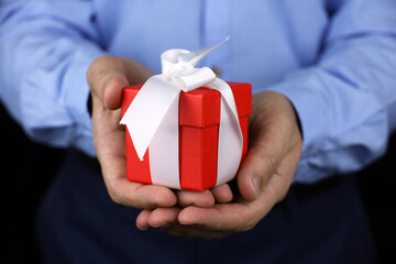 Red gift box in male hands close up. Man in office clothes giving present with white ribbon, concept of Christmas, birthday or Valentine's day