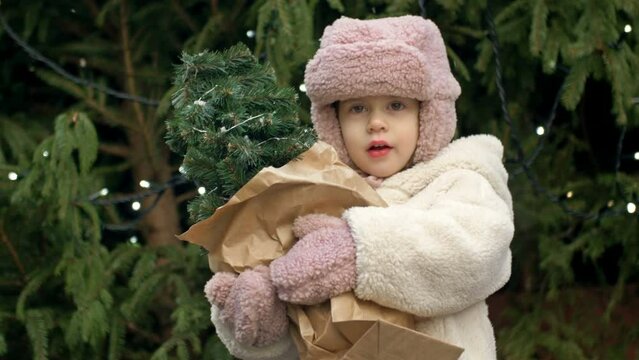 A 2-3 year old girl at the Christmas market holding a small Christmas tree. A child catches the falling snowflakes with her tongue.