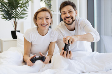 Front view of concentrated caucasian couple playing video games while lying under blanket at home. Smiling man and woman looking on tv have fun competing in video game using joysticks.