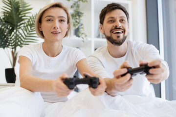 Fototapeta premium Close up of concentrated caucasian couple playing video games while lying under blanket at home. Smiling man and woman looking on tv have fun competing in video game using joysticks.