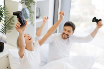 Happy woman with raising hands with joystick looking at husband celebrating victory in video game. Side view of caucasian couple in love looking at tv have fun competing in video game on bed.