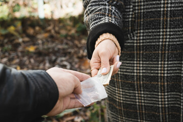 Male drug dealer sells hard drugs in a transparent plastic bag to an addicted woman with money in her hand, cropped image - obrazy, fototapety, plakaty