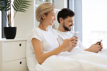 Serene caucasian family of two using mobile phones while lazing in soft bed during daytime at home. Confident adult man and woman reading online article paying attention to details.