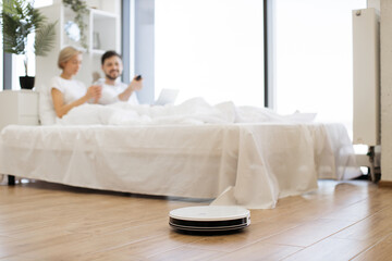 Selective view of modern robot vacuum cleaning wooden floor in bedroom while young multiracial couple watching video on laptop. Young family saving time for leisure by using electrical device. - obrazy, fototapety, plakaty