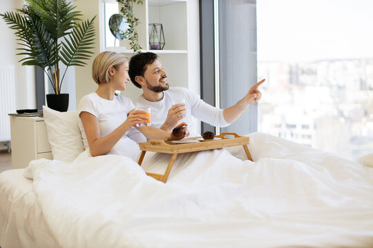 Caucasian family man and woman eating muffins and drinking orange juice while lying on bed in morning. Joyful married couple looking at panoramic window sending message of true love through daily care