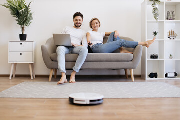 Positive caucasian man and woman sitting together on sofa and using remote controller for robot vacuum cleaner. Concept of smart home functions and fully automated gadget. - obrazy, fototapety, plakaty