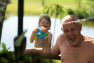 Kid girl and grandfather playing with water gun toy in the summer.