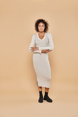 brunette african american woman standing in midi dress and boots on beige backdrop, autumn style
