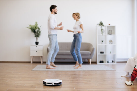 Young caucasian lady dancing with male partner while robotic vacuum running floor smoothly. Homeowners using electrical appliance for household chores while freeing up time for mother-daughter party.