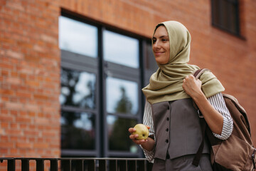 Beautiful woman in hijab standing on city street. Muslim studnet eating apple, going to school....