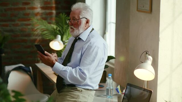 Old businessman in full suit sitting on table in office looking at smartphone,chatting online, sending messages in office late night