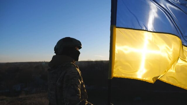 Ukrainian army girl stands with lifted national banner against background of sunset. Young female soldier in military uniform holds a waving flag of Ukraine to peak of hill. Victory of war. Dolly shot