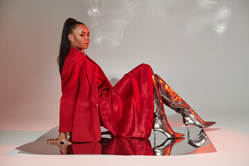 young african american woman in red dress, blazer and silver boots sitting on mirrored surface